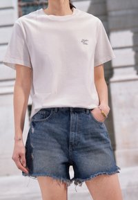 Osoba v bílém tričku s textem "Crazy Sexy Cool" a vysokými roztrhnutými denimovými šortkami, stojící venku s rukama v kapsách.