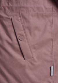 Tissu imperméable mauve avec une poche à rabat cousue sécurisée par un bouton et une petite étiquette "WATERPROOF" sous la poche.