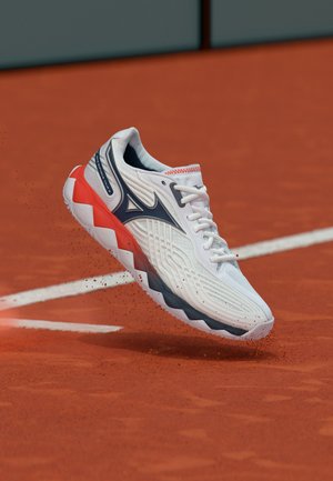 Chaussure de sport blanche, noire et rouge flottant au-dessus d'un court de tennis en terre battue avec des marques blanches, soulevant de petites particules d'argile.