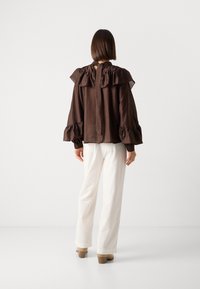 Brun bluse med rynket krave og manchetter, rygknaplukning, parret med hvide ribbede vidde bukser og beige blokhælede sko.