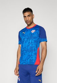 Maglia sportiva a maniche corte blu e rossa con un motivo testurizzato, dotata di collo a giro e logo Puma. Indossata con pantaloni corti blu coordinati.