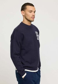 Sweatshirt en tricot marine avec un col rond, texte blanc "LON DON CITI" sur la poitrine, poignets et ourlet côtelés ; coupe décontractée avec épaules tombantes.