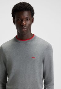 Grey knit sweater with a round neckline and red accent. Features a small red logo on the chest and textured fabric.