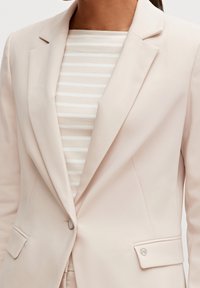 Jasnoróżowa marynarka o gładkiej fakturze, z zapięciem na pojedynczy guzik i kieszeniami naszytymi, nałożona na miękki, paskowany beżowy top.