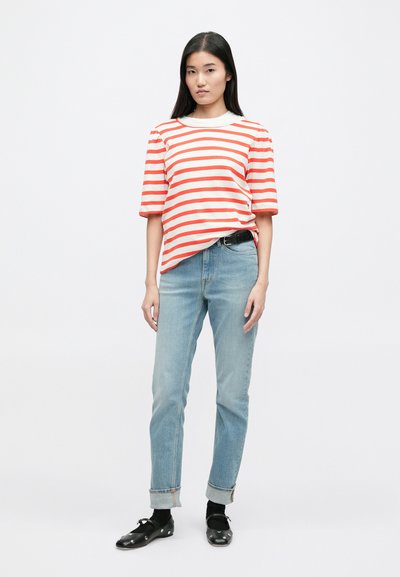 Κόκκινο και άσπρο ριγέ κοντομάνικο πουκάμισο, χαλαρή εφαρμογή, σε συνδυασμό με ανοιχτό γαλάζιο τζιν, γυρισμένα στους αστραγάλους, και μαύρα παπούτσια με λευκές λεπτομέρειες.