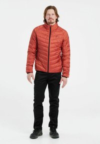 Giacca rossa trapuntata con collo alto, chiusura frontale con zip e dettagli neri. Indossata con pantaloni neri e calzature scure. Design minimalista.