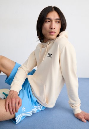 Kremna kapuca s črnim logotipom Adidas je v kombinaciji z modrimi kratkimi hlačami, ki imajo bele črte. Mehka tkanina in sproščen kroj. Postavljena na modri površini.