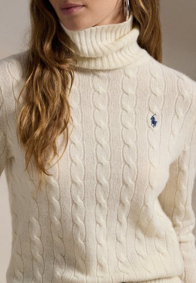 Personne aux cheveux longs portant un pull à col roulé en maille torsadée crème avec le logo Polo brodé en bleu et une boucle d’oreille argentée en forme d’anneau.