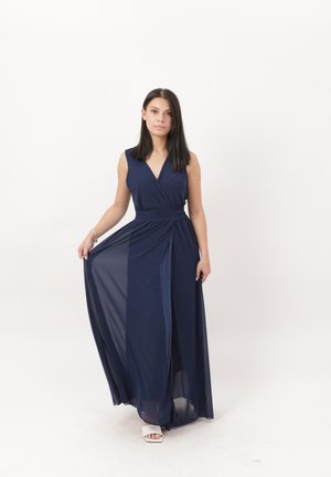 Vrouw met stijl donker haar die een mouwloze marineblauwe maxijurk en witte sandalen draagt, terwijl ze de jurk rok iets opzij houdt.
