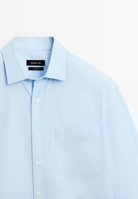 Camicia azzurra chiara con bottoni, realizzata in tessuto liscio, caratterizzata da un colletto classico, taschino sul petto e bottoni bianchi. Design semplice e pulito.