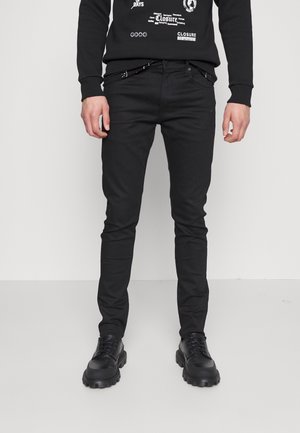 Mann trägt enge schwarze Jeans, schwarzen Sweatshirt mit Grafik und schwarze klobige Schnürschuhe, steht vor weißem Hintergrund.