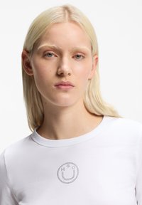 Ξανθιά γυναίκα με ανοιχτόχρωμο δέρμα που φοράει ένα λευκό πουκάμισο με σχέδιο μιας χαμογελαστής φατσούλας με τελείες στο στήθος, πάνω σε ένα απλό λευκό φόντο.