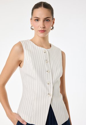 Kobieta nosząca białą, bezrękawną bluzkę w wąskie paski z guzikami oraz srebrne kolczyki koła, stojąca na jednolitym białym tle.