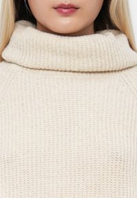 Cremefarbenes Strickpullover mit einem dicken Rollkragen. Der Stoff wirkt weich und strukturiert und weist ein grobes Rippmuster auf.