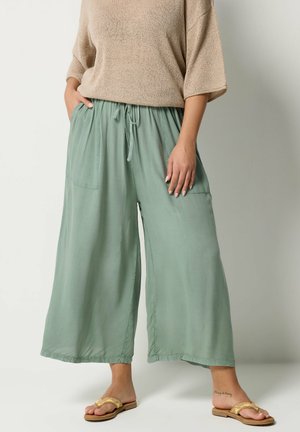Person iført brede lysegrønne bukser med forlommer, beige kortærmet top og gule sandaler, stående med hænderne i lommerne.