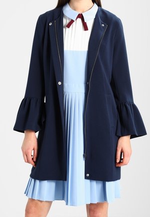 Femme portant une veste bleu marine à manches évasées sur une robe plissée bleu clair avec un col blanc et un ruban rouge.