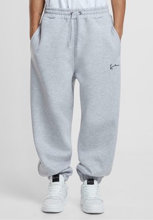 Homme portant un pantalon de survêtement gris clair avec des poignets élastiques et une taille à cordon, associé à des baskets blanches sur un fond uni.