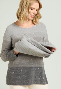 Grijze trui met een verloopontwerp, voorzien van geribbelde manchetten en verspreide versieringen, vastgehouden door een persoon. Zachte textuur en een losse pasvorm.