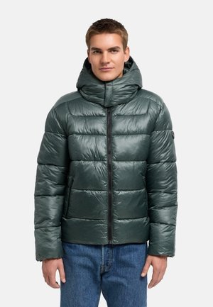 Giovane uomo con giacca imbottita verde scuro con collo alto e cappuccio, abbinata a jeans blu, in piedi davanti a uno sfondo semplice.