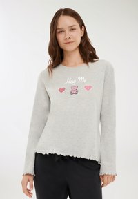 Giovane donna che indossa un top a maniche lunghe grigio chiaro con bordi scallop e la scritta "Hug Me", cuori rosa e un design di orsetto.
