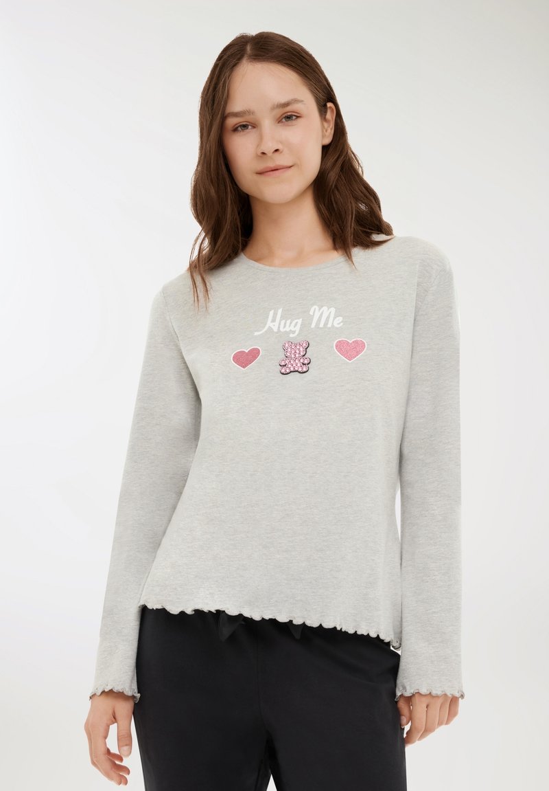 Giovane donna che indossa un top a maniche lunghe grigio chiaro con bordi scallop e la scritta "Hug Me", cuori rosa e un design di orsetto.
