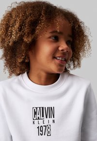 Felpa bianca con scollatura rotonda e tessuto texturizzato, caratterizzata da un logo nero prominente che reca la scritta "CALVIN KLEIN 1978" in lettere grasse.