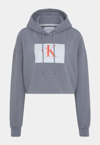 Szara krótka bluza z kapturem, z długimi rękawami, przednimi sznurkami do regulacji oraz białą prostokątną łatą z logo, na której widnieją napisy "cK" i "Calvin Klein Jeans" w kolorze czerwonym i białym.