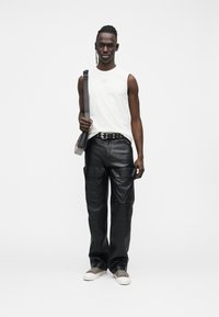 Wit mouwloos shirt, zwarte leren cargobroek met zakken, grijze sneakers en een crossbodytas. Zwarte riem met zilveren accenten.