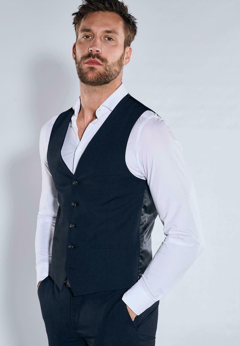 Marineblaues Weste mit elegantem Design, ausgestattet mit einem satinierten Rücken, fünf Knöpfen und Seitentaschen. Getragen über einem weißen Hemd.