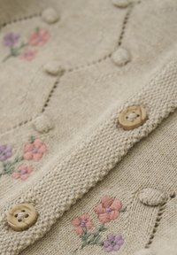 Virkat kofta i beige med blommor broderade i rosa och lila. Har träknappar och en strukturerad tyg med upphöjda detaljer.