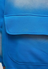 Blauwe sweater met een grote, gevouwen voorzak. Het materiaal is zacht, met een gladde textuur en een verloop van licht naar donkerblauw.