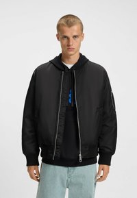 Veste bomber noire avec une texture lisse, dotée d'une fermeture éclair, de poignets côtelés et de deux poches latérales. En dessous, un hoodie noir est visible.