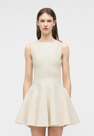 Γυναίκα που φοράει αμάνικο, ελεφαντόδοντο φόρεμα με υφή, φούστα με φάκιδα και διακοσμητικό κλιπ στον αριστερό ώμο, στέκεται μπροστά σε απλό φόντο.