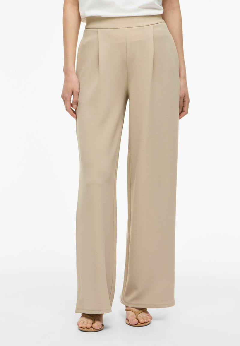 Pantaloni beige a gamba larga con pieghe anteriori indossati con sandali sabbia e top bianco infilato, mostrati dalla vita ai piedi.