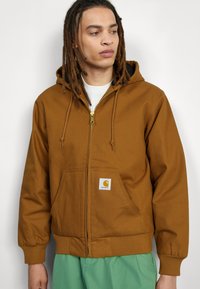 Brown zip-up hoodie with a drawstring hood, front pockets, ribbed cuffs, and hem. Includes a small Carhartt logo patch on the front.