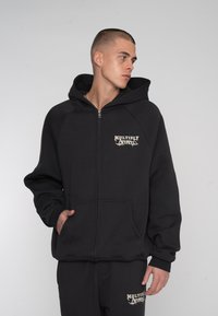 Schwarze Zip-Hoodie aus weichem Material. Verfügt über eine Fronttasche und ein Logo auf der Brust. Dieses Modell hat eine Kapuze und gerippte Bündchen.