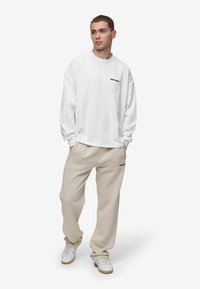 Sudadera blanca de manga larga con texto negro, combinada con pantalones de chándal beige. El conjunto tiene un ajuste relajado y un diseño sencillo.