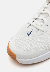 Biele sieťované atlétske topánky s bielymi šnúrkami a modrým logom Nike; majú gumenú podrážku a textúrovaný zvršok pre priedušnosť.