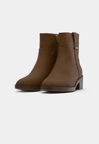 Καφέ ψεύτικες δερμάτινες μπότες έως τον αστράγαλο με φερμουάρ στο πλάι, χαμηλό τακούνι και λεία υφή. Απλός σχεδιασμός χωρίς ορατά σχέδια ή διακοσμητικά στοιχεία.