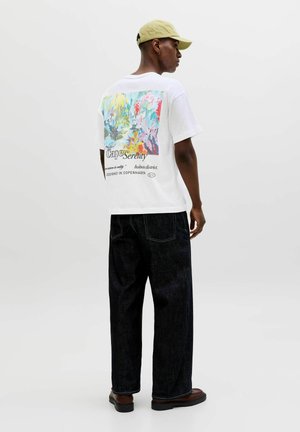 Uomo che indossa una T-shirt bianca con grafica floreale colorata e testo sul retro, jeans neri a gamba larga, scarpe marroni e un berretto beige.