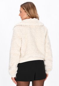 Kvinde med skulderlangt hår iført en hvid, struktureret fleecejakke, sorte shorts og øreringe, vist fra ryggen og i sideprofil.