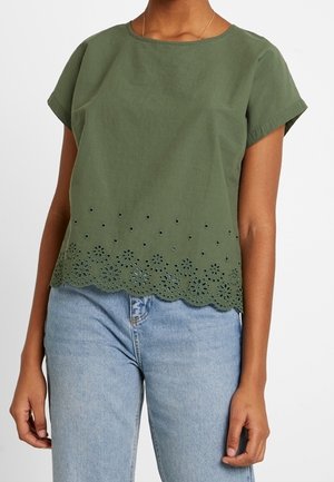 Vrouw draagt een olijfgroene blouse met korte mouwen en bloemvormige uitgesneden patronen aan de zoom, gecombineerd met lichtblauwe jeans.