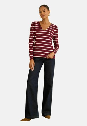 Kastanienbraunes und cremefarbenes gestreiftes Langarmoberteil mit einem gewellten Ausschnitt, kombiniert mit dunklen Jeanshose im Schlag und beigen Hausschuhen.