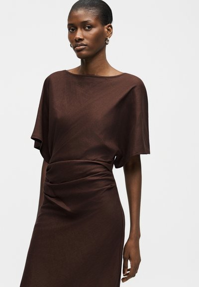 Γυναίκα που φοράει σκούρο καφέ φόρεμα με κοντά μανίκια, άνετο πάνω μέρος και εφαρμοστή, ελαφρώς σούρες μέση, στέκεται μπροστά σε απλό ανοιχτό φόντο.