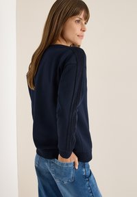 Marineblå sweatshirt med strukturerede sidepaneler, ribbede manchetter og en afslappet pasform. Båret sammen med slidte blå jeans, vist fra en sidevinkel.