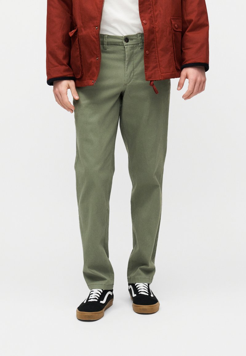 Άνδρας που φοράει χακί παντελόνι, κόκκινο σακάκι, λευκό πουκάμισο και μαύρα αθλητικά παπούτσια με λευκά κορδόνια, στεκόμενος μπροστά σε απλό φόντο.