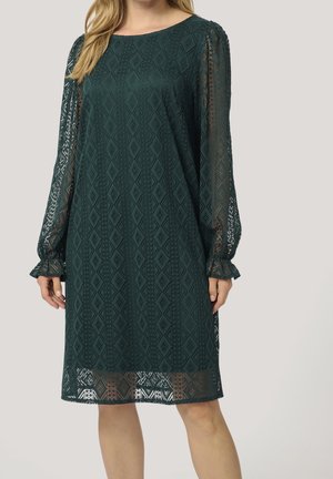 Frau, die ein dunkelgrünes Spitzenkleid mit langen durchsichtigen Ärmeln und knielangem Saum trägt, mit einem geometrischen Diamantmuster.