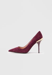 Γυναικεία μπορντό σουέτ γόβα με ψηλό τακούνι και μυτερή μύτη, διακοσμημένη με χρυσή λεπτομέρεια κοντά στον τακούνι, με φόντο μια απλή ανοιχτόχρωμη επιφάνεια.