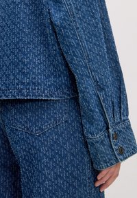 Blaues Jeansjacke mit strukturiertem Muster, langen Ärmeln und Knopfbündchen. Der Stoff ist robust und hat einen leicht verwitterten Look.
