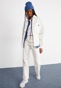 White denim jacket over a blue denim shirt, paired with white jeans and gray sneakers. A blue knitted beanie completes the look.
